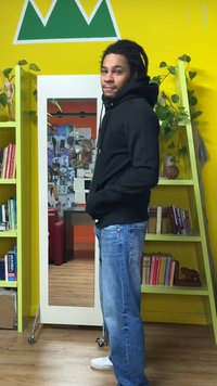 Camisola com capuz preta com bolso frontal, usada com jeans azuis e sapatilhas brancas. Em uma sala iluminada com decoração verde e amarela, reflexo espelhado visível.