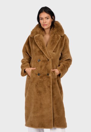 Femme portant un long manteau en fausse fourrure brune avec un grand col et les mains dans les poches, se tenant devant un fond clair.