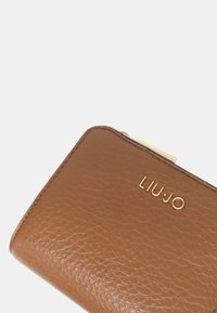 Portafoglio in pelle marrone con superficie texture, logo dorato e cuciture sottili lungo i bordi. Forma rettangolare compatta.