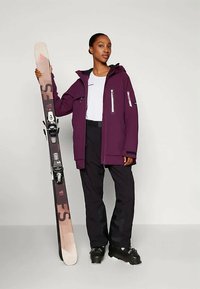 Egy nő lila síkabátot, fekete nadrágot és síbakancsot visel, és egyetlen rózsaszín-fekete hegyi sílécet tart függőlegesen egy sima háttér előtt.