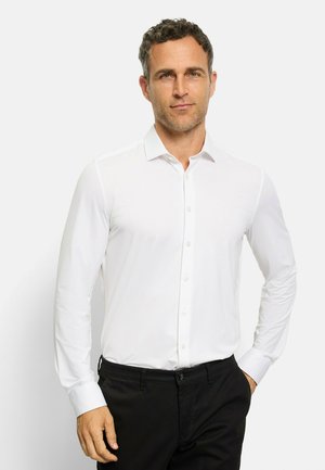Camicia bianca a maniche lunghe con colletto classico, apertura con bottoni e texture liscia. Indossata con pantaloni neri, che mostrano un design aderente.