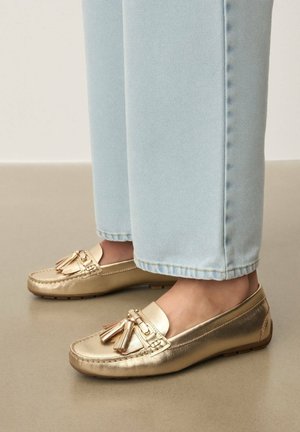 Fényes arany színű bojtos mokaszinban álló lábak, világoskék egyenes szárú farmerral, sima padlón.