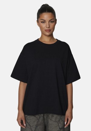 Donna con i capelli scuri raccolti in uno chignon, indossa una camicia nera oversize a maniche corte e pantaloni con motivo pietra, in piedi davanti a uno sfondo bianco uniforme.