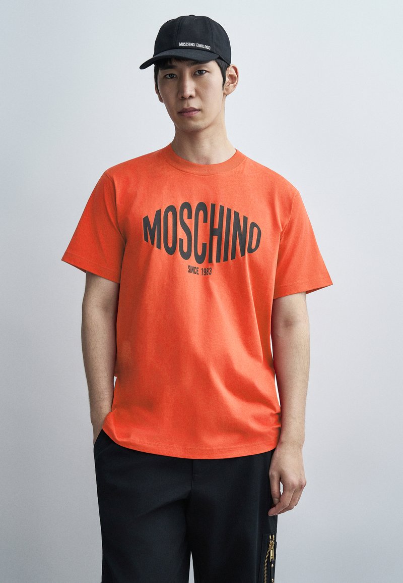 Πορτοκαλί βαμβακερό μπλουζάκι με μαύρο κείμενο "MOSCHINO" και έτος 1983. Συνδυάζεται με μαύρο σορτς με φερμουάρ. Το καπέλο είναι μαύρο, με το λογότυπο της μάρκας.