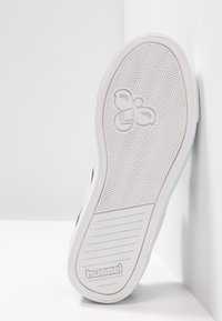 White rubber outsole with herringbone pattern, featuring a logo imprint. The shoe has a flat shape and is angled against a white wall.