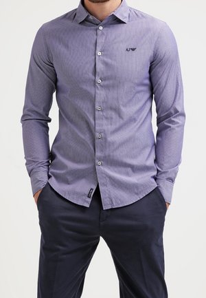 Man draagt een slim-fit, blauw gestreepte, lange mouw overhemd met kraag en donkere broek, handen in de zakken.