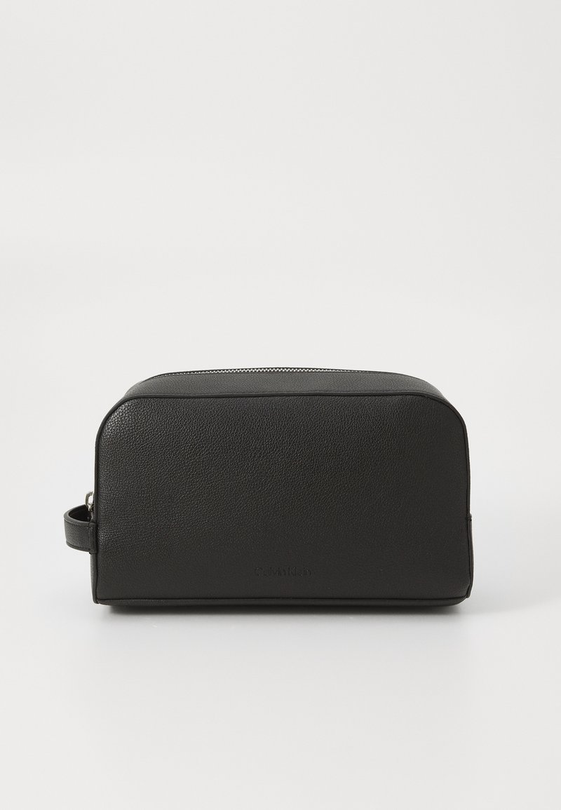 Trousse de toilette en cuir noir de forme rectangulaire, surface texturée, fermeture zippée sur le dessus et poignée latérale pour un transport facile.