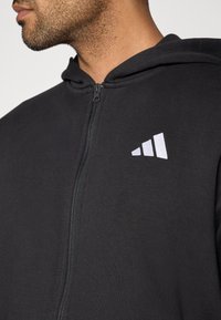 Čierna mikina na zips z bavlneného zmesi materiálu, ktorá má podšitú kapucňu, predný zips a výrazné biele logo na hrudi.