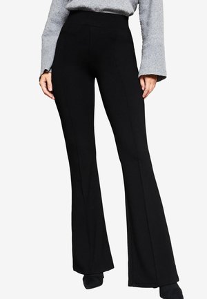Vrouw draagt zwarte hoge taille flare broek en een grijze gebreide trui met wijde mouwen, staand tegen een effen achtergrond.
