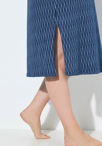 Abito midi blu navy con strisce bianche verticali, dotato di uno spacco laterale. Tessuto liscio con una leggera lucentezza. A piedi nudi su una superficie chiara.
