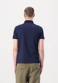 Marineblått polo-skjorte med et subtilt mønster, kort erm, og en kontrasterende mørk krage. Brukes med olivengrønne bukser, som viser en glattet tekstur.