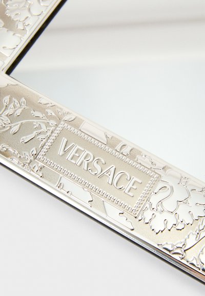 Sudraba krāsas taisnstūra formas spoguļa rāmis ar izsmalcinātiem ziedu rakstiem un centrā iespiestu "VERSACE" logotipu. Gluda metāliska tekstūra.