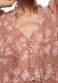 Braun-blühende Bluse mit pinken und weißen Mustern, einem gerafften Ausschnitt und Knopfleiste vorne. Weiches Material mit leichter Textur.