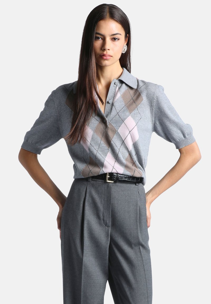 Jeune femme aux longs cheveux foncés portant un pull gris à manches courtes à motif argyle et un pantalon gris taille haute avec une ceinture noire.