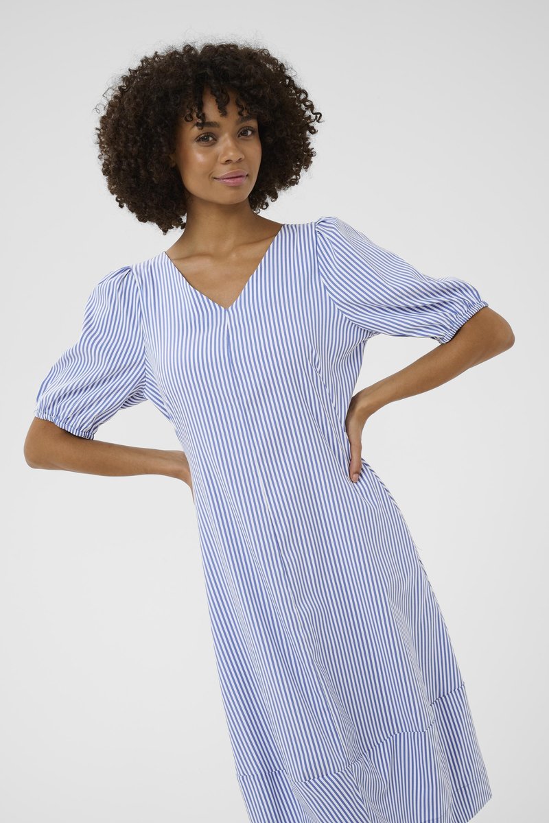 Lokkidega naine, kes kannab sinise ja valge vertikaalsete triipudega kleiti lühikeste volangvarrukatega, positsioneerides käed puusadel halli tausta ees.