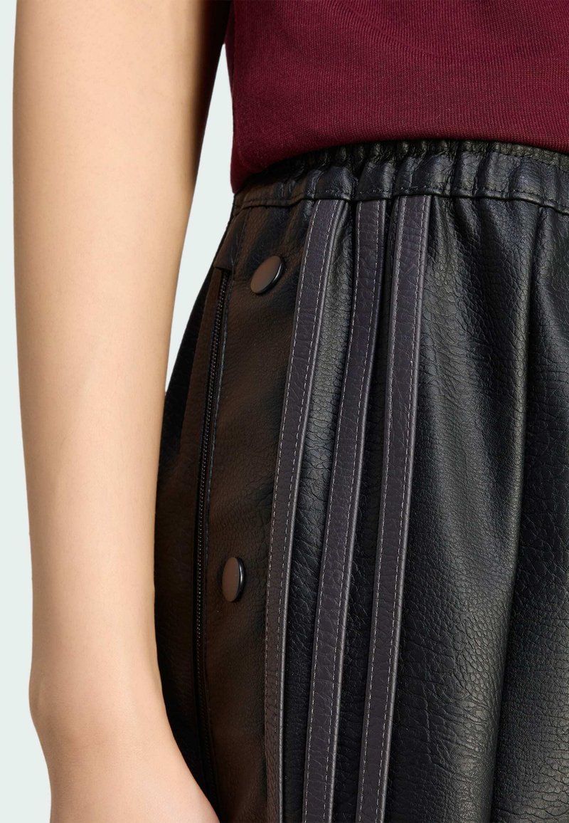 Close-up of hand beside black textured leather pants with snap buttons and vertical stitched stripes, paired with a dark red top.