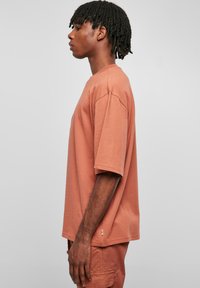 Tricou scurt de culoare ruginită, confecționat din bumbac moale, având o croială relaxată și o siluetă pătrată. Suprafață texturată cu cusături vizibile.