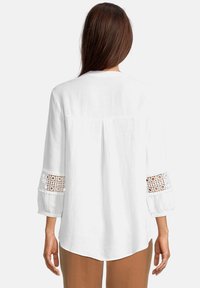 Blusa bianca a maniche lunghe con dettagli in pizzo ai polsi, texture morbida e vestibilità rilassata; presenta una scollatura rotonda e dettaglio di piega sul retro.