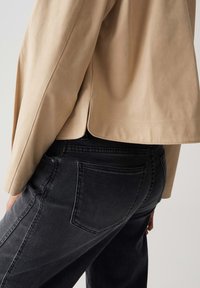 Giacca in suede beige con orlo curvo, indossata sopra jeans in denim neri con una tasca posteriore e una leggera texture.