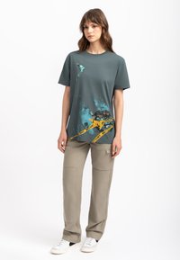 Grijze t-shirt met grafisch ontwerp van verfspatten en "Natte verf" waarschuwingslint; gecombineerd met beige cargo-broek en witte sneakers.