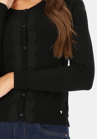 Czarny kardigan z teksturową koronkową wstawką z przodu, zapinany na pięć guzików, z prążkowanymi mankietami. Subtelny akcent logo na dolnym brzegu.