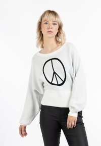 Suéter gris claro con un símbolo de paz negro, escote ancho y dobladillo acanalado. Las mangas tienen forma de globo y están ajustadas en el puño.