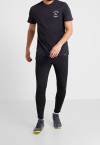 Zwarte t-shirt met witte opdruk "Ici C'est Paris", gecombineerd met zwarte leggings en grijze voetbalschoenen met gele accenten.
