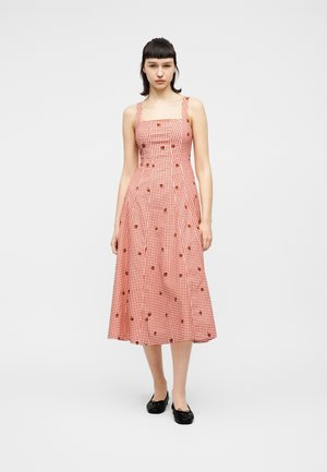 Kvinde står iført en ærmeløs midikjole med rød- og hvide tern og små blomsterprint samt sorte ballerinasko.