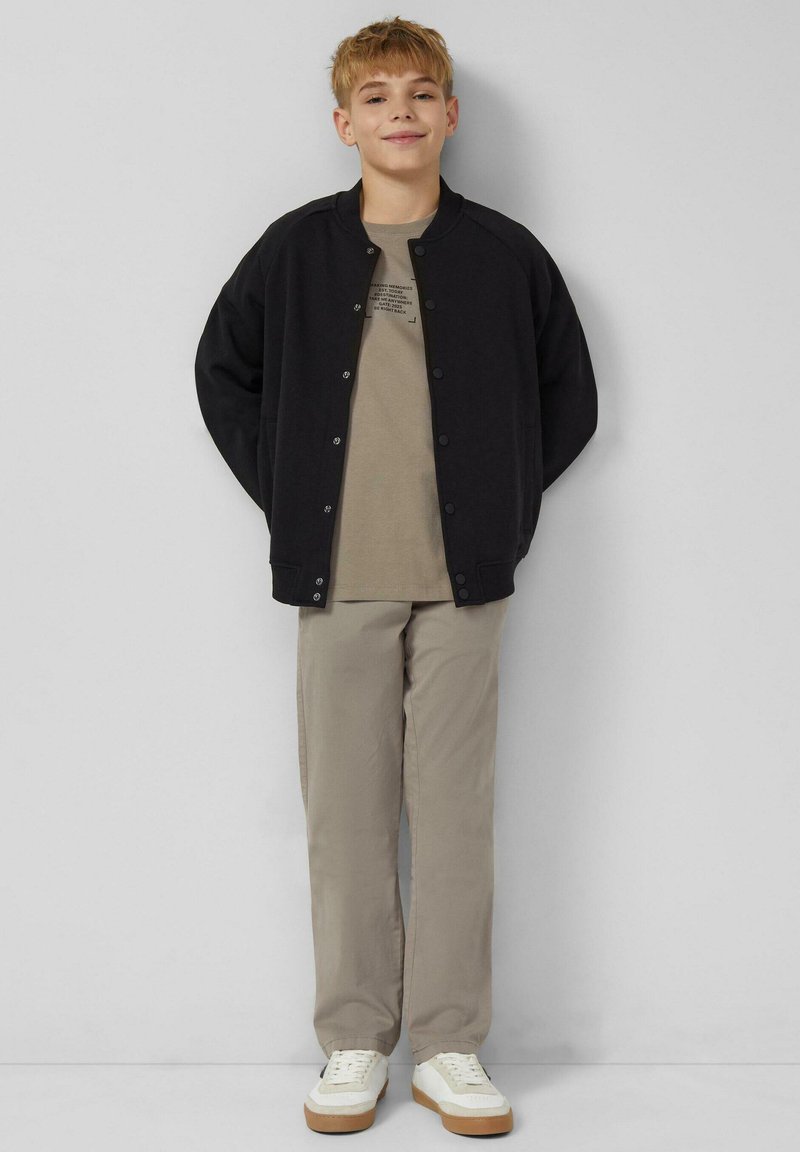 Czarna kurtka bomber, beżowy t-shirt z nadrukiem, khaki spodnie i białe sneakersy z gumową podeszwą. Casualowy, luźny krój całego stroju.