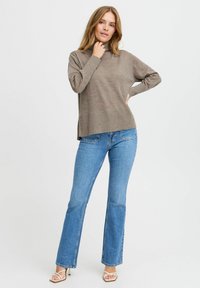 Pull en laine marron avec un col montant, coupe décontractée et fentes sur les côtés, associé à un jean évasé bleu clair et des talons à lanières.
