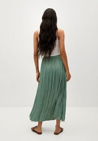 Vrouw met lang donker haar draagt een witte mouwloze top, een groene geplooide midi-rok en bruine slidesandalen, staande tegen een witte achtergrond.