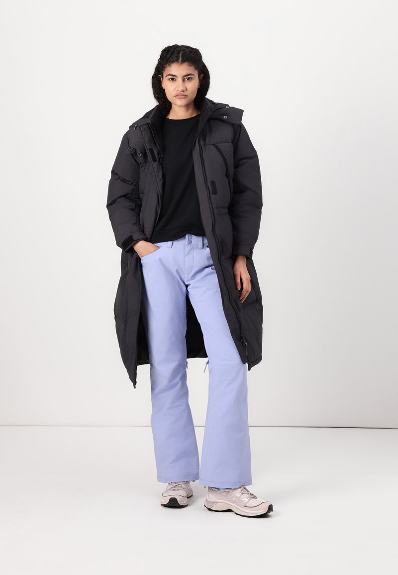 Schwarze gepolsterte Jacke mit Kapuze, Fronttaschen, kombiniert mit hellblauen Hosen und hellen Sneakern. Glatte Textur und oversize Schnitt.