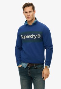 Niebieska bluza z kontrastowym pasem w kolorze granatowym i zielonym, z logo "Superdry". Połączona z dżinsową koszulą i ciemnymi dżinsami.