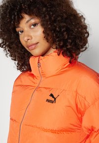 Giacca puffer PUMA in un vivace arancione, con colletto alto, zip frontale completa e logo nero sul petto. Texture liscia e lucida.