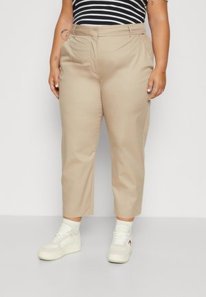 Person wearing beige cropped trousers, white sneakers, white socks, and a black and white striped shirt, standing indoors against a plain background.