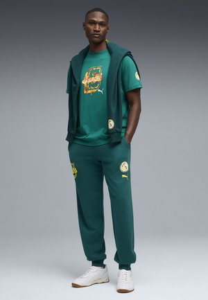 Mand iført grøn Puma-træningsdragt med Senegal-tema og hvide sneakers, stående med hænderne i lommerne mod en ensfarvet grå baggrund.