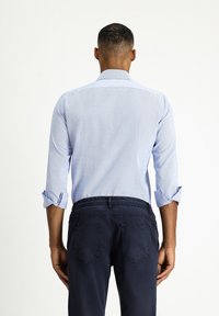 Lichtblauw, geknoopd shirt met opgerolde mouwen, met een subtiel patroon. Gecombineerd met donkerblauwe broek, zicht van achter.