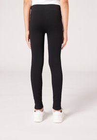 Leggings negras con textura acanalada y cinturilla elástica, de longitud ajustada hasta el tobillo, combinadas con zapatillas blancas, con una textura suave y sin patrones visibles.
