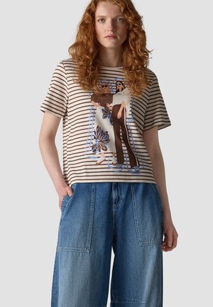 Young woman with curly red hair wearing a brown and white striped shirt with graphic of two fashionably dressed women and wide blue denim pants.