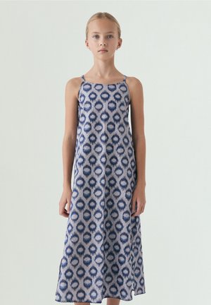 Niña joven con cabello rubio con un vestido largo sin mangas de patrones azules y blancos, de pie frente a un fondo liso.