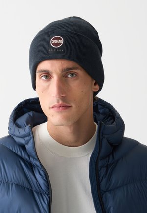 Sötétkék kötött sapka bordázott textúrával, kerek logóval, amelyen az áll: "COLMAR ORIGINALS". Kék steppelt dzsekivel és fehér pulóverrel viselve.