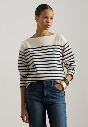Krémový top s dlhým rukávom s námorníckymi horizontálnymi pruhmi, širokým výstrihom, voľného strihu, v kombinácii s modrými džínsami a masívnym náramkom.