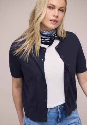 Donna bionda che indossa un cardigan blu navy a maniche corte con bottoni, camicia bianca, jeans blu e sciarpa a righe al collo, su uno sfondo semplice.