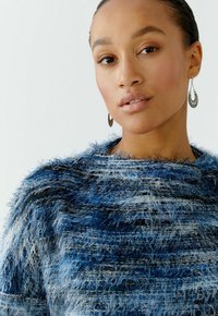Blau-weißer strukturierter Pullover mit einem flauschigen, mehrschichtigen Design. Er hat einen weiten Ausschnitt und ein Modell trägt einen runden Ohrring.