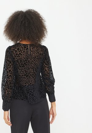 Mujer con cabello rizado que lleva una blusa negra translúcida de manga larga con estampado de leopardo y pantalones negros, de pie de espaldas al espectador.