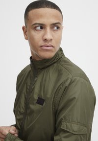 Olive green jacket with a high collar, zip closure, and black accent patch on the chest. Features a smooth, shiny texture and cuff details.