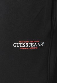 Svart tyg med en slät textur har en framträdande logotyp i vit och röd text: "AMERICAN TRADITION GUESS JEANS ORIGINAL DESIGNS."