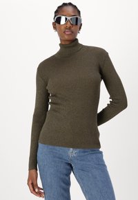 Olive green ribbed turtleneck sweater in a fitted design, paired with light blue jeans and mirrored sunglasses.