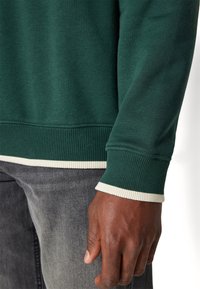 Närbild av en hand och handled som bär en mörkgrön tröja med vit kant, tillsammans med mörkgrå jeans.