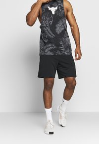 Czarny top bez rękawów z białym logo byka i wzorem liści, w połączeniu z czarnymi szortami i jasnymi butami sportowymi. Charakteryzuje się wygodnym krojem i sportowym designem.